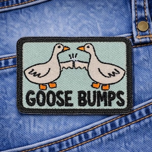 May include: Embroidered patch featuring two cartoon geese bumping beaks, with the words "GOOSE BUMPS" in black lettering. The patch has a light blue background and a black border. The patch is on a blue denim background.