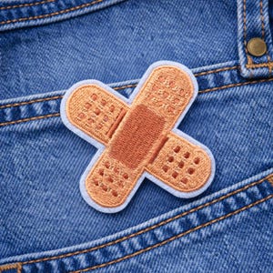 May include: Embroidered patch in the shape of a bandage, with a light blue border. The bandage is tan and brown, with a detailed stitched design. The patch is placed on a blue denim background.