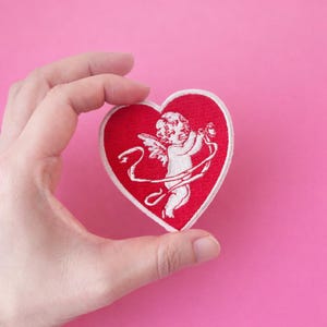 May include: A red heart-shaped patch with a white embroidered image of Cupid holding a bow and arrow. The patch has a white border and is set against a pink background.