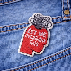 May include: Embroidered patch with the text "LET ME OVERTHINK THIS" on a red head-shaped design. The patch has a white border and a gray, tangled brain graphic. The patch is placed on a blue denim background.