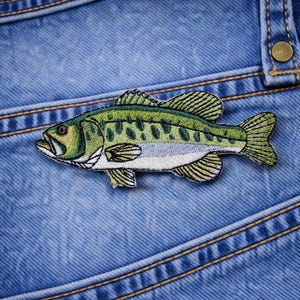 May include: Embroidered patch of a green and white bass fish on a blue denim background. The fish has detailed scales and fins. The denim has visible stitching and a pocket detail.