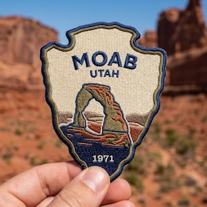 May include: A shield-shaped embroidered patch featuring a beige background with a dark blue border. The text MOAB UTAH is stitched in dark blue above a depiction of a natural stone arch. The number 1971 is stitched in white at the bottom.