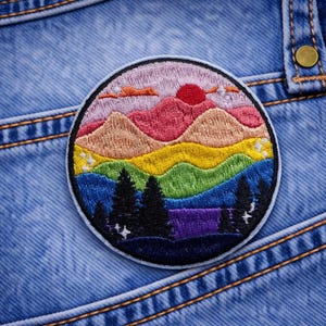 May include: Embroidered patch featuring a colorful mountain landscape with a red sun, layered mountains, and trees. The patch is round with a dark blue border and is set against a blue denim background.