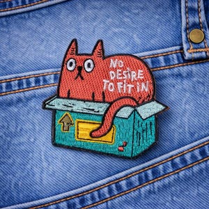 May include: Embroidered patch featuring a red cat sitting in a teal box. The patch reads "NO DESIRE TO FIT IN" in white text. The patch is placed on a blue denim background.