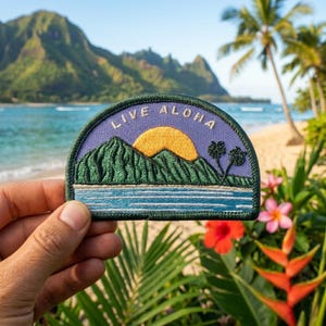 May include: Embroidered patch with a tropical scene featuring a mountain range, ocean, and a setting sun. The patch is in shades of green, blue, and yellow, with the words "LIVE ALOHA" in white lettering. The patch is held in front of a beach scene.
