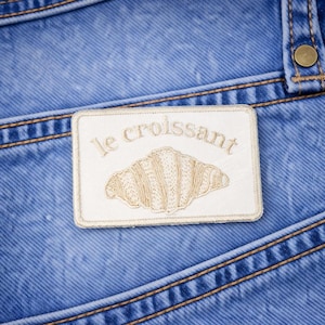 May include: Close-up of blue denim jeans with a rectangular patch. The patch features the words "le croissant" above an embroidered croissant design. The jeans have visible stitching and a button.