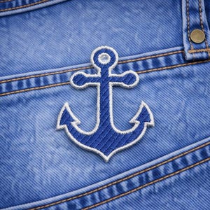May include: Close-up of blue denim fabric with a stitched blue and white anchor patch. The anchor is centered, with a detailed design. The denim has visible seams and a button.