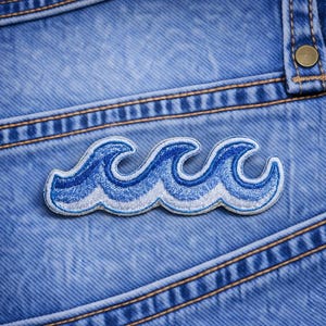 May include: A blue and white embroidered wave patch on a denim background. The patch features three stylized waves with a light blue and white color scheme. The denim has visible stitching and a button.
