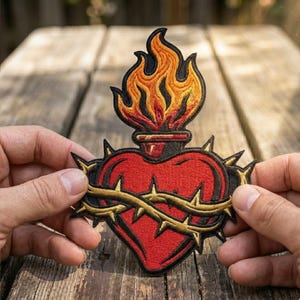 May include: Embroidered patch featuring a red heart with a gold and black crown of thorns. Above the heart is a flame design in shades of orange and yellow. The patch is held against a wooden background.