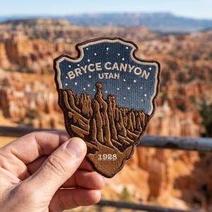 May include: A shield-shaped embroidered patch featuring a brown landscape of Bryce Canyon, Utah, against a dark blue starry sky. The text BRYCE CANYON UTAH is stitched in white at the top, with the year 1928 embroidered in white at the bottom.
