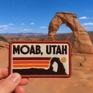 May include: Embroidered patch featuring the text "MOAB, UTAH" above a stylized landscape with a sunset design and the silhouette of an arch. The patch has a rectangular shape with a brown border.