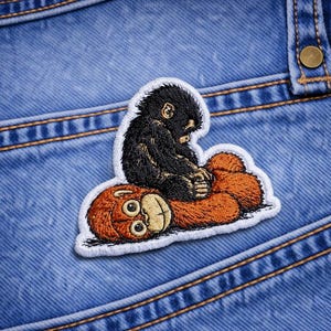 May include: Embroidered patch featuring a black monkey sitting on a brown orangutan. The patch has a white border and is set against a blue denim background. The patch is a fun, whimsical design.