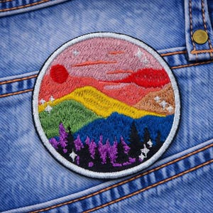 May include: A round embroidered patch featuring a colorful mountain landscape with a red sun and pink sky. The patch includes green, yellow, blue, and purple trees and mountains, with a white border. The patch is set against a blue denim background.