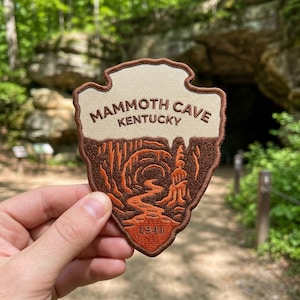 May include: A shield-shaped embroidered patch featuring a cream-colored top section with the brown text MAMMOTH CAVE KENTUCKY. The bottom section depicts a brown and orange cave interior scene with the year 1941 printed at the very base.