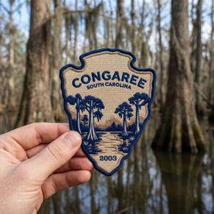 May include: A shield-shaped embroidered patch featuring a tan background with a dark blue border. The text reads CONGAREE SOUTH CAROLINA and 2003. The design depicts a swamp landscape with dark blue cypress trees and a winding waterway.