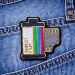 May include: Embroidered patch featuring a film roll design with the text "SHOOT FILM." The patch has a black border, a white and colorful striped center, and a gray film strip detail. It is placed on a blue denim background.