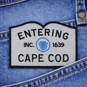 May include: A rectangular patch with the text "ENTERING INC. CAPE COD 1639" in black lettering, with a blue emblem. The patch is set against a blue denim background, with visible stitching.