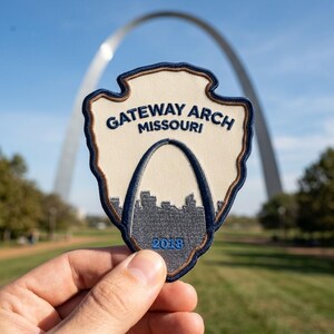 May include: A shield-shaped embroidered patch featuring a cream-colored background with dark blue text reading GATEWAY ARCH MISSOURI and the year 2018. The patch displays a dark blue outline of the Gateway Arch monument over a gray city skyline.