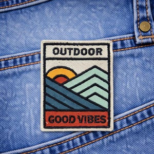 May include: Embroidered patch with the word "OUTDOOR" at the top and "GOOD VIBES" at the bottom. The patch features a mountain landscape with a setting sun in orange, red, and blue tones. The patch is rectangular and set on a blue denim background.