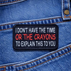 May include: Black rectangular patch with white text reading "I DON'T HAVE THE TIME OR THE CRAYONS TO EXPLAIN THIS TO YOU" in a bold font. The text "OR THE CRAYONS" is in red. The patch is sewn onto a blue denim background.