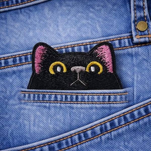 May include: A black cat patch with pink inner ears and yellow eyes peeks out of a blue jean pocket. The patch is embroidered and has a detailed design, with the jeans' stitching visible.