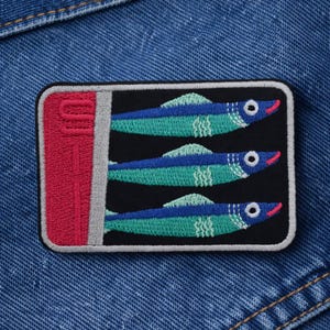 May include: Embroidered patch featuring three stylized sardines in a can. The sardines are blue and teal with red accents, set against a black background. The can is red with a silver pull tab, all on a denim background.
