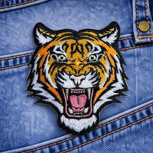 May include: Embroidered tiger head patch with orange, black, white, and yellow details. The tiger has an open mouth, revealing pink and white teeth. The patch is on a blue denim background.