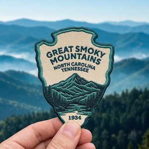 May include: An embroidered patch shaped like an arrowhead with a cream background and dark teal border. It features the text GREAT SMOKY MOUNTAINS NORTH CAROLINA TENNESSEE and 1934, with a dark teal graphic of mountain peaks and a river.