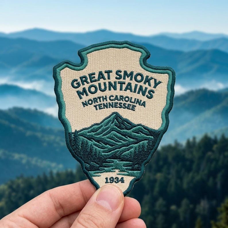 May include: An embroidered patch shaped like an arrowhead with a cream background and dark teal border. It features the text GREAT SMOKY MOUNTAINS NORTH CAROLINA TENNESSEE and 1934, with a dark teal graphic of mountain peaks and a river.