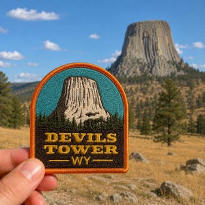 May include: A patch featuring Devils Tower in Wyoming. The patch has a brown border, teal sky, and a detailed image of the tower. The text "DEVILS TOWER - WY-" is in gold lettering. The background shows a natural landscape with a blue sky.
