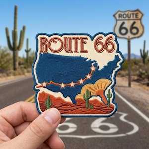 May include: An embroidered patch featuring a dark blue map of the United States with a curved line of white stars, a desert landscape with green cacti, and the text ROUTE 66 in brown lettering against a cream background with a dark blue border.