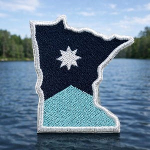 May include: Embroidered patch in the shape of the state of Minnesota. The patch features a navy blue upper section with a white star and a light blue lower section. The patch has a white border.