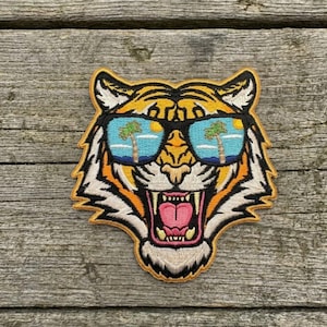 May include: Embroidered patch featuring a tiger head wearing sunglasses. The sunglasses reflect a beach scene with palm trees and a setting sun. The tiger has orange, black, and white stripes, with a pink tongue and open mouth.