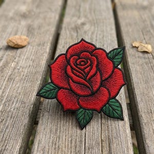 May include: Embroidered red rose patch with green leaves and a black outline. The rose is in full bloom, showcasing detailed stitching. This patch is suitable for jackets, bags, or other fabric items.