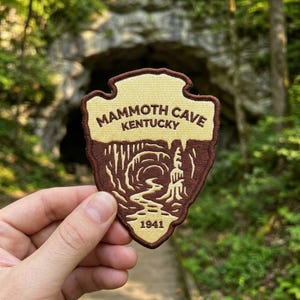 May include: An embroidered patch shaped like a shield with a dark brown border. The top section is cream-colored with the text MAMMOTH CAVE KENTUCKY. The bottom section is brown with a graphic of a cave interior and the year 1941 at the base.