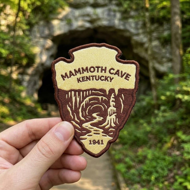 May include: An embroidered patch shaped like a shield with a dark brown border. The top section is cream-colored with the text MAMMOTH CAVE KENTUCKY. The bottom section is brown with a graphic of a cave interior and the year 1941 at the base.