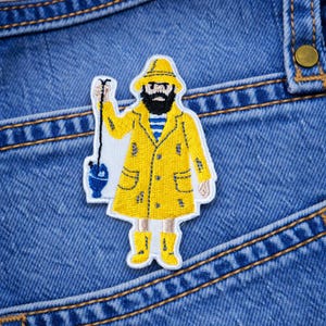 May include: Embroidered patch featuring a cartoon fisherman in a yellow raincoat, hat, and boots. The fisherman has a black beard and is holding a fishing rod with a blue bucket. The patch is on a blue denim background.