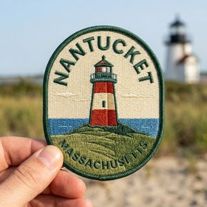 May include: An oval embroidered patch featuring a red and white lighthouse on a grassy hill with blue water. The text NANTUCKET is arched above the lighthouse, and MASSACHUSETTS is written in a curve at the bottom, all bordered by dark green.