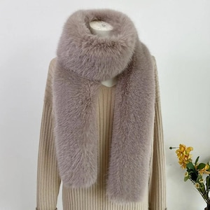 May include: A luxurious, light brown faux fur scarf displayed on a mannequin. The scarf has a wide, fluffy collar and long, draping ends. The soft, textured material is a neutral color, suitable for various outfits. The scarf is a winter accessory.