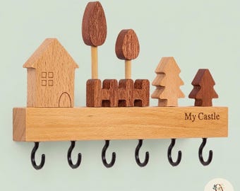 May include: A wall-mounted wooden key holder featuring a light wood base with the text My Castle engraved on the right side. The top displays a house, a fence, and four trees in varying shades of brown. Six black metal hooks hang from the base.