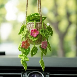 Op de afbeelding: Een handgemaakte gehaakte plantenbak met een beige pot, groene bladeren en roze en oranje bloemen. De plantenbak wordt opgehangen met een beige touw, wat een decoratief en grillig tintje geeft. Het haakwerk is gedetailleerd.