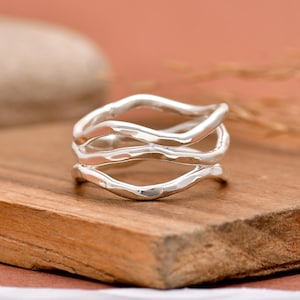 May include: A silver ring featuring a multi-layered design with three distinct, wavy, and interconnected bands. The metal surface has a polished, reflective finish with organic, flowing curves that create an open, airy structure throughout the piece.