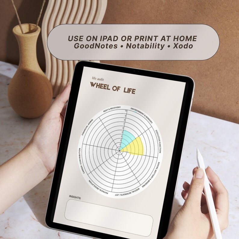 A digital Wheel of Life self-assessment displayed on a tablet. The wheel is divided into segments with labels like "Rest & Recovery" and "Environment & Sensory." The text "USE ON IPAD OR PRINT AT HOME" is at the top. A stylus is held near the screen.