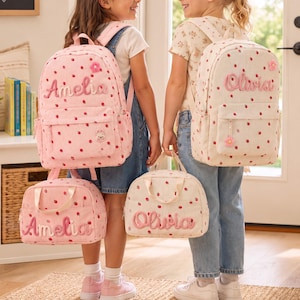 May include: Two personalized backpacks and two matching lunch bags featuring a pink and white strawberry pattern. One set is embroidered with the name Amelia, and the other set is embroidered with the name Olivia, both in a cursive script font.