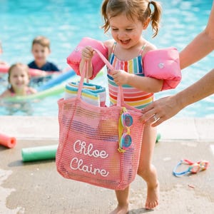May include: A pink mesh beach bag with the name Chloe Claire embroidered in white script on the front. The bag features two handles and a pair of colourful swimming goggles attached to the side, containing a folded striped towel inside.