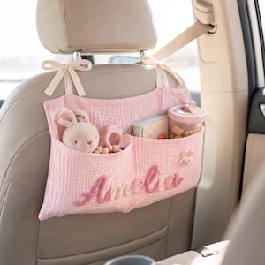 Pode incluir: Um organizador de assento de carro em musselina rosa claro com dois bolsos, preso ao encosto de cabeça com fitas creme. O nome Amelia está bordado em rosa na frente, acompanhado por um pequeno aplique de coelho, um peluche e um copo.