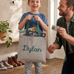 May include: A grey and white pinstriped fabric tote bag with green handles and the name Dylan embroidered in green script on the front. The bag contains two books titled Dinosaur Dig and Space Explorer, and it is held by a smiling child.