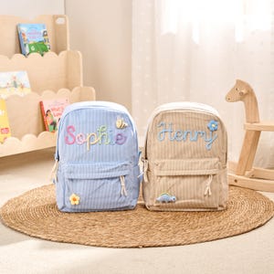 May include: Two corduroy backpacks, one light blue and one beige, sit on a woven mat. The blue backpack has the name "Sophie" embroidered in rainbow thread, and the beige backpack has the name "Henry" embroidered in blue thread. Each backpack has a front pocket and decorative patches.