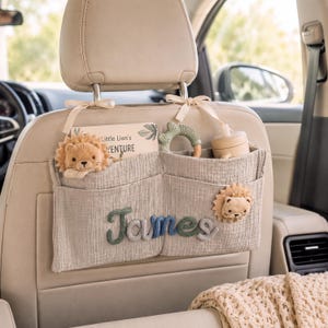May include: A light gray muslin car seat organizer featuring the name James in blue and green yarn lettering. The organizer has two pockets containing a book titled Little Lion's Adventure, a green silicone ring, and a beige cup with a lid.