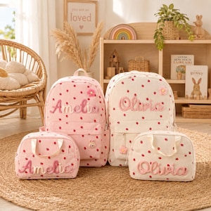 May include: Two corduroy backpacks and two matching lunch bags featuring a red strawberry pattern. One set is light pink with the name Amelia embroidered in pink, and the other is cream with the name Olivia embroidered in pink.